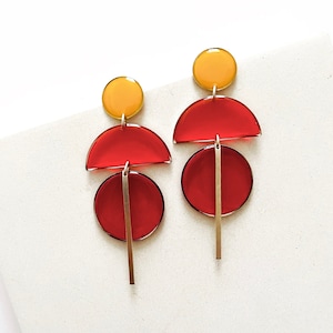 May include: A pair of geometric earrings with red and yellow resin pieces. The earrings feature a circle, a half-circle, and a long, thin bar.