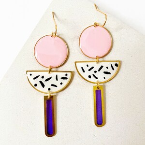Puede incluir: Un par de pendientes geom&eacute;tricos con esmalte rosa, blanco y morado. Los pendientes tienen un c&iacute;rculo grande, un semic&iacute;rculo y un rect&aacute;ngulo largo.