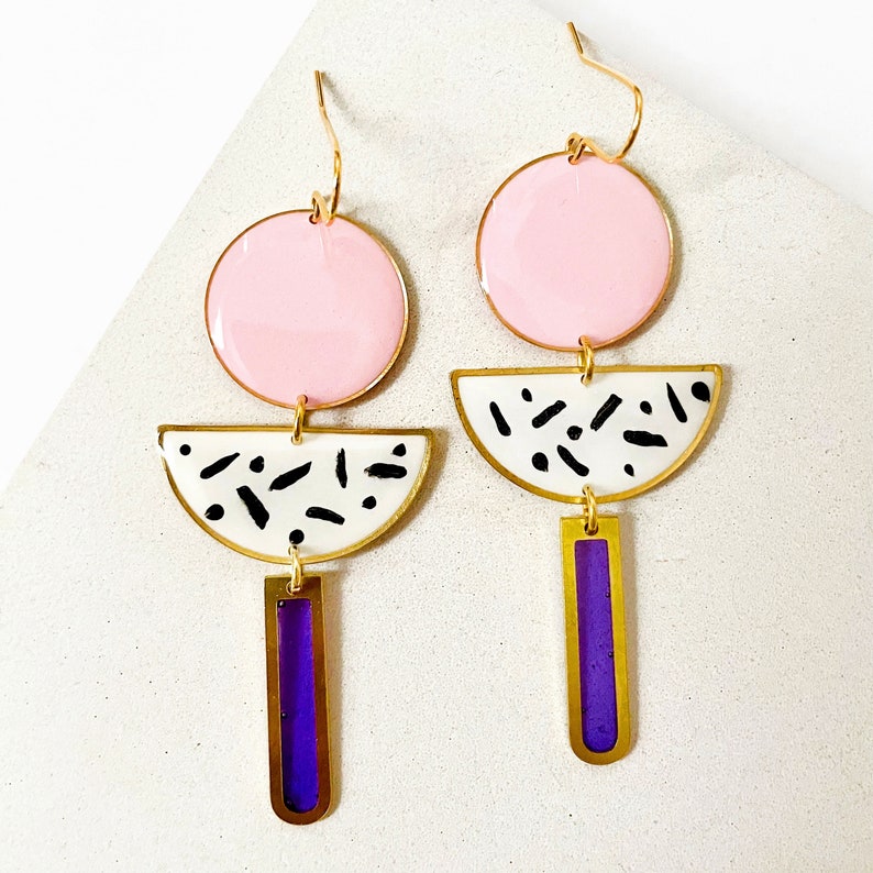 Puede incluir: Un par de pendientes geom&eacute;tricos con esmalte rosa, blanco y morado. Los pendientes tienen un c&iacute;rculo grande, un semic&iacute;rculo y un rect&aacute;ngulo largo.