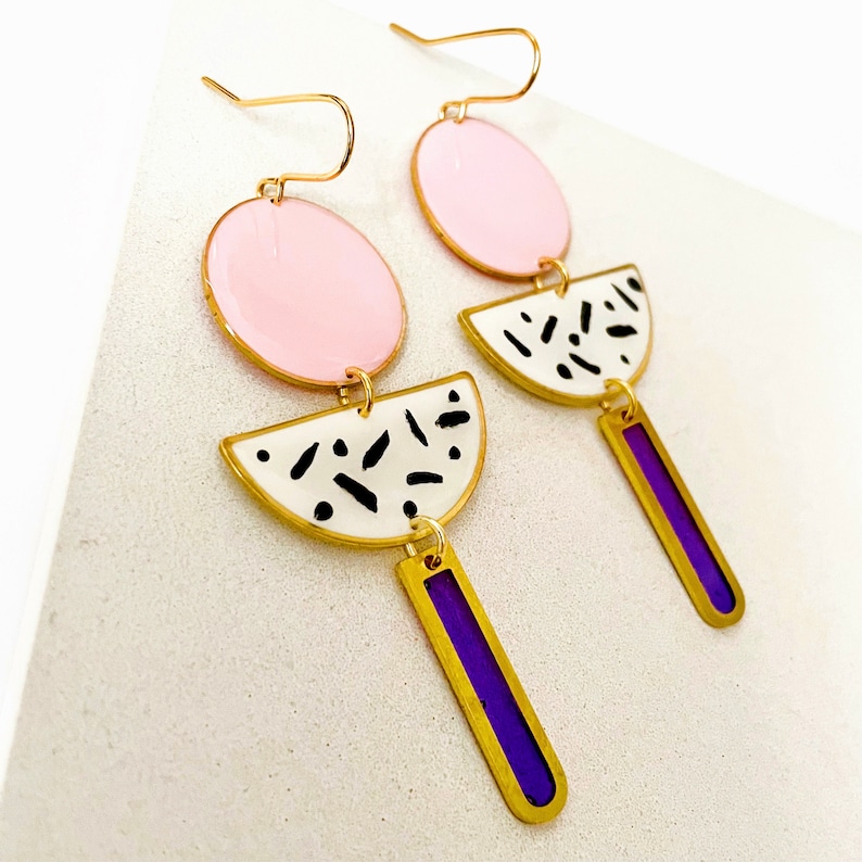 Puede incluir: Un par de pendientes colgantes con un dise&ntilde;o geom&eacute;trico rosa, blanco y negro. Los pendientes presentan un c&iacute;rculo rosa, un semic&iacute;rculo blanco y negro y un rect&aacute;ngulo morado.