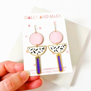 Puede incluir: Un par de pendientes geom&eacute;tricos con esmalte rosa, blanco y negro. Los pendientes tienen forma de c&iacute;rculo, semic&iacute;rculo y rect&aacute;ngulo. Los pendientes est&aacute;n en una tarjeta blanca con el texto "DALLY AND MACK" y una direcci&oacute;n de sitio web.