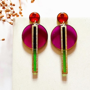 Puede incluir: Un par de pendientes geométricos con un gran disco redondo rosa y rojo y una larga barra verde y negra que cuelga debajo. Los pendientes están hechos de metal dorado.