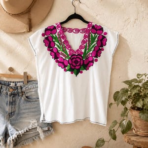 May include: White embroidered top with a vibrant floral design in shades of pink and green. The top is sleeveless and has a V-neckline. A pair of denim shorts and a straw hat are also visible in the image.