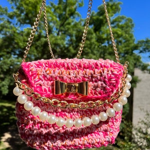 May include: A small pink crocheted handbag with a gold chain strap and pearl trim. The bag has a gold bow closure.