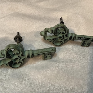 May include: Two decorative key-shaped cabinet pulls with an antique finish. The pulls are a distressed teal color with black accents, featuring ornate scrollwork and keyhole details. Each pull has a metal screw for mounting.