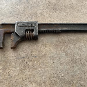 May include: An antique adjustable wrench, featuring the "Ford USA" logo. The wrench is made of dark, aged metal with visible rust, indicating its vintage. The adjustable jaw is open, and the handle is long and slender.