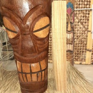 May include: Two wooden tiki statues, one with a carved face and the other with a more abstract design. Both statues are brown and have a weathered look. The statues are standing on a straw mat.