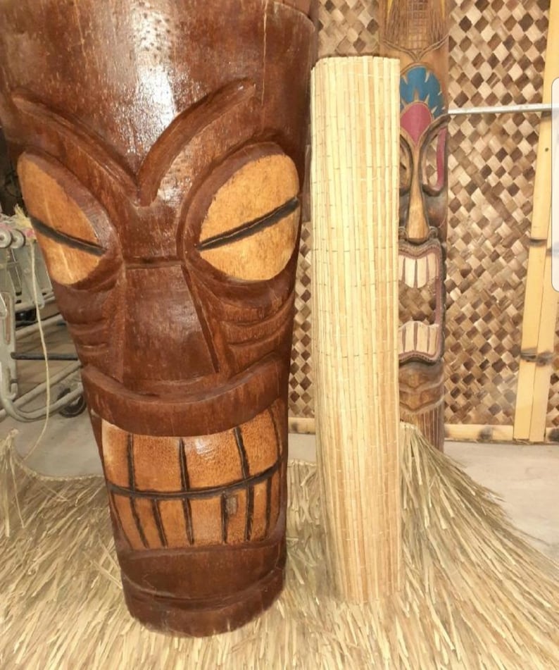 May include: Two wooden tiki statues, one with a carved face and the other with a more abstract design. Both statues are brown and have a weathered look. The statues are standing on a straw mat.