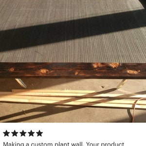 May include: A partially constructed wooden frame with dark stained edges and a gray slatted interior. The frame is resting on a concrete surface, with tools and lumber visible in the background. The text says, "Making a custom plant wall. Your product worked great."