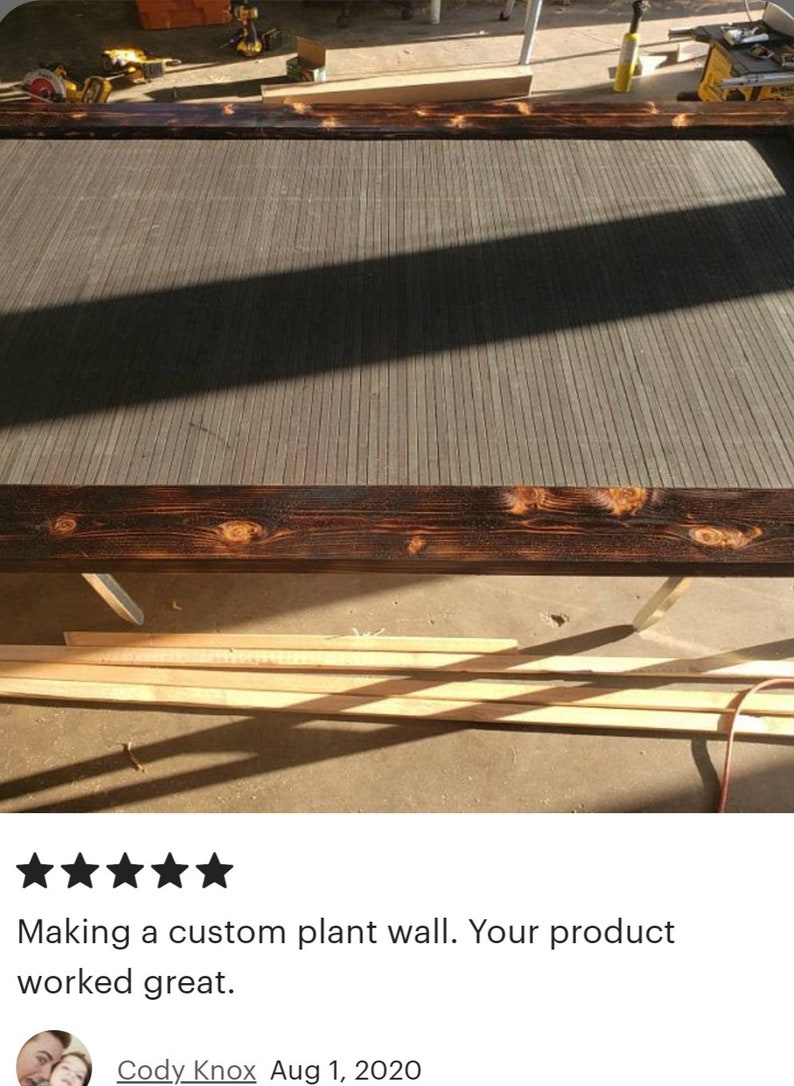 May include: A partially constructed wooden frame with dark stained edges and a gray slatted interior. The frame is resting on a concrete surface, with tools and lumber visible in the background. The text says, "Making a custom plant wall. Your product worked great."