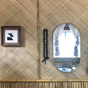 May include: A framed black and white print of a surfer riding a wave hangs on a wall with a woven bamboo pattern. A black beaded necklace hangs on the wall to the right of the print. A large oval mirror with a wooden frame reflects a window with white curtains.
