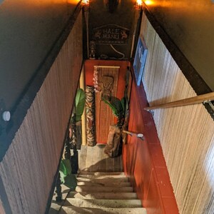 May include: A wooden staircase with a tiki-themed decor, featuring a carved wooden tiki head, a sign that reads "Hale Manu", and a bamboo-like wall covering.