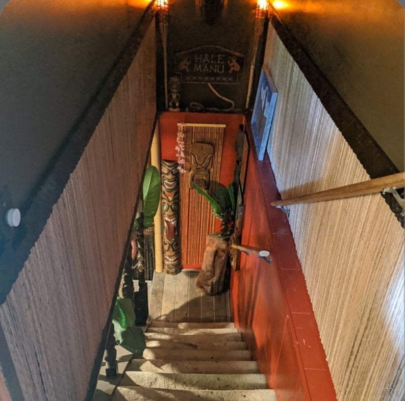 May include: A wooden staircase with a tiki-themed decor, featuring a carved wooden tiki head, a sign that reads "Hale Manu", and a bamboo-like wall covering.