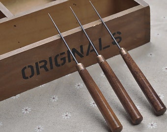 May include: Three wooden-handled tools with metal tips. The tools are arranged in a row on a light brown fabric surface. The tools are in a wooden box with the word "ORIGINALS" printed on the side.