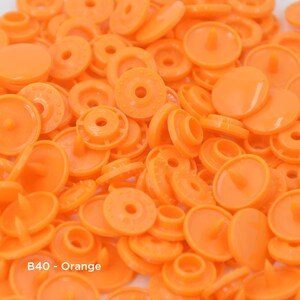 May include: A close-up of a pile of orange plastic snap fasteners. The fasteners are round and have a small, raised center. The text "B40 - Orange" is visible in the lower left corner of the image.