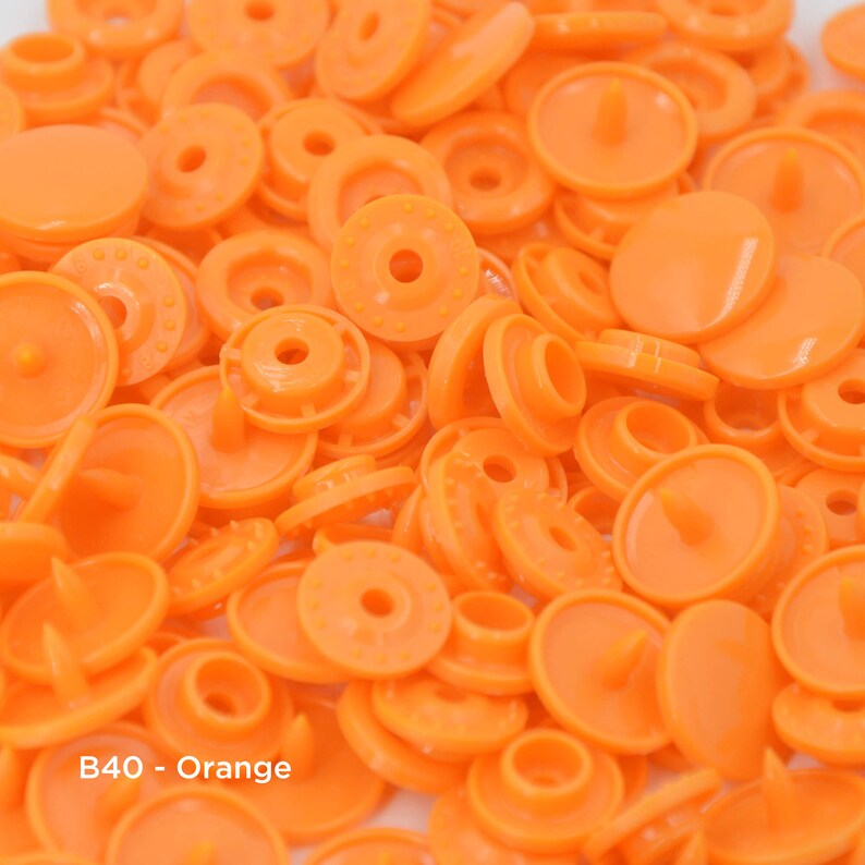 May include: A close-up of a pile of orange plastic snap fasteners. The fasteners are round and have a small, raised center. The text "B40 - Orange" is visible in the lower left corner of the image.