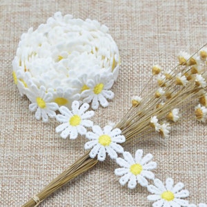 Puede incluir: Borde de encaje blanco con flores de margarita amarillas. El borde está enrollado y colocado sobre un fondo de tela marrón.