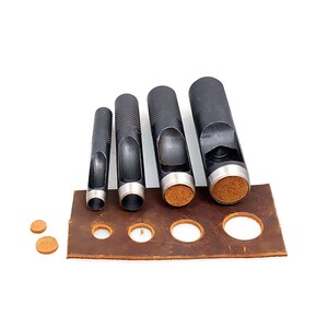 May include: Set of five black leather hole punch tools with different sized round holes. The tools are arranged in order of size, with the largest tool on the right and the smallest tool on the left. The tools are sitting on a brown leather surface with a few leather circles.