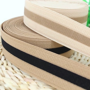 Puede incluir: Dos rollos de elástico beige con rayas negras. El elástico está tejido con un patrón texturizado.