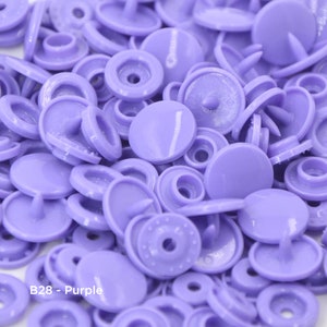 May include: A close-up view of a large quantity of purple plastic snap buttons. The buttons are a light shade of purple and have a smooth, shiny finish. The buttons are arranged in a random pattern, with some buttons stacked on top of each other. The text "B28 - Purple" is visible in the lower left corner of the image.