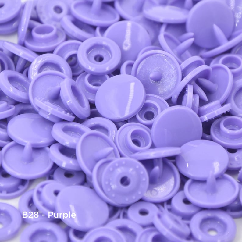 May include: A close-up view of a large quantity of purple plastic snap buttons. The buttons are a light shade of purple and have a smooth, shiny finish. The buttons are arranged in a random pattern, with some buttons stacked on top of each other. The text "B28 - Purple" is visible in the lower left corner of the image.