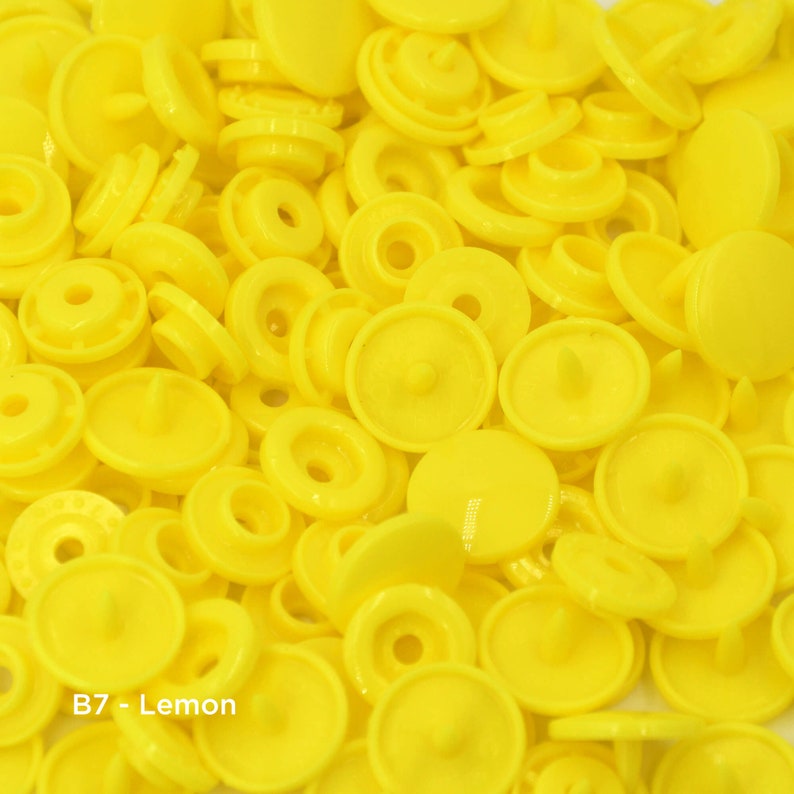 May include: A close-up image of a large quantity of bright yellow plastic snap buttons. The buttons are in various sizes and shapes, with some having a raised center and others having a flat surface. The text "B7 - Lemon" is visible in the lower left corner of the image.