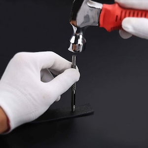 May include: A hand wearing a white glove holds a leather punch tool while another hand uses a red and silver hammer to punch a hole in a piece of black leather.