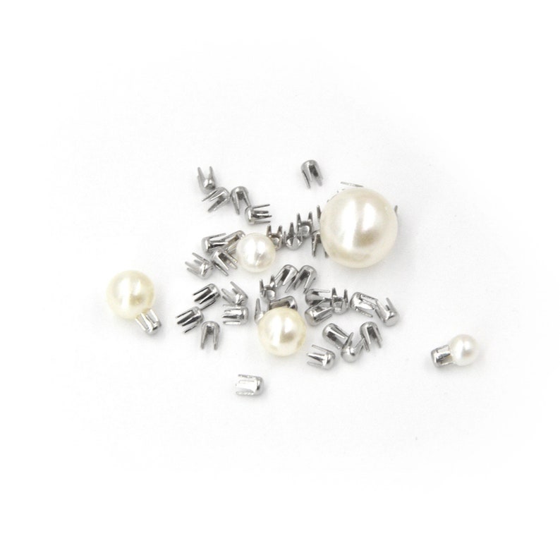 May include: A collection of silver metal nail heads and pearl-like beads, scattered on a white surface.