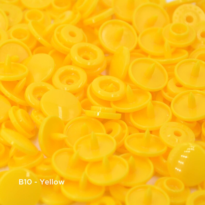 May include: A close-up image of a pile of bright yellow plastic snap buttons. The buttons are all the same size and shape, and they have a small, raised center.
