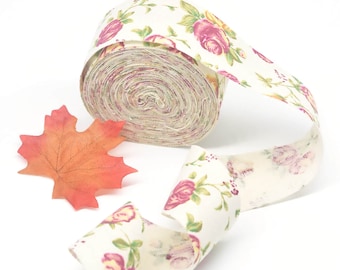 May include: A roll of white fabric ribbon with a floral print featuring pink roses and green leaves. The ribbon is rolled up and a portion of it is unrolled, showing the pattern.
