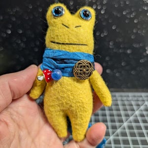 May include: A handmade yellow felt frog doll with large blue eyes and a blue scarf. The frog has a decorative button and a red and white polka dot accent. The doll is held in a person's hand.