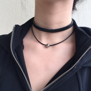 Könnte beinhalten: Eine schwarze Lederkette mit einem silbernen Perlenakzent. Die Kette ist mit einer zweiten, dünneren schwarzen Kette übereinander gelegt.