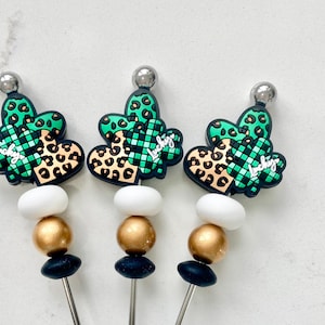 以下が含まれることがあります： 黒、金、白のビーズが付いたステンレス製のカクテルピックが3本。各ピックの先端には、緑と茶色のハート型のチャームが付いており、緑のクローバーと「Lucky」という文字が印刷されています。