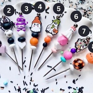 Peut inclure: Huit cake pops sur le thème d'Halloween avec différents motifs, dont un fantôme, une sorcière, une momie, une araignée et une citrouille. Chaque cake pop est décoré de perles colorées et d'une brochette en métal.