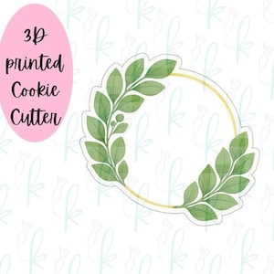 Puede incluir: Cortador de galletas impreso en 3D con forma de corona de hojas verdes y contorno dorado. El texto "3D printed Cookie Cutter" está en negro sobre un óvalo rosa.