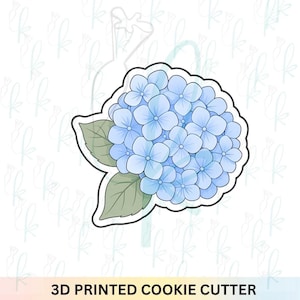 May include: A 3D printed cookie cutter shaped like a hydrangea flower cluster. The design features a rounded head of light blue petals with two pale green leaves extending from the side, outlined in a thick white border against a light background.