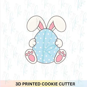 Peut inclure: Emporte-pièce imprimé en 3D représentant un lapin tenant un œuf de Pâques bleu à pois. Le lapin a des oreilles blanches avec l'intérieur rose et des coussinets roses. L'œuf est orné d'un nœud. Le texte dit : 3D PRINTED COOKIE CUTTER.