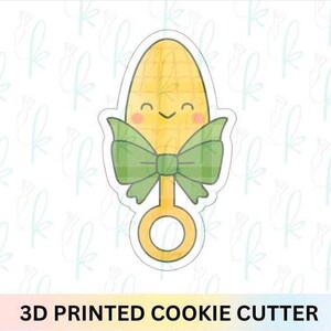 May include: A 3D printed cookie cutter shaped like a smiling corn cob, featuring a green plaid bow and a ring handle. The text "3D PRINTED COOKIE CUTTER" is at the bottom.
