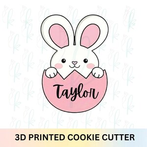 Könnte beinhalten: Ein weißer Hasen-Ausstecher mit rosa Innenohren und Wangen, der aus einem rosa Ei kommt. Der Name "Taylor" ist in schwarzer Schrift auf dem Ei geschrieben. Der Text "3D PRINTED COOKIE CUTTER" steht unten.