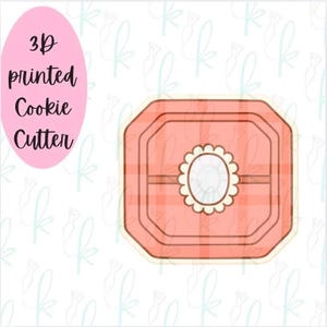 May include: A square cookie cutter with rounded corners and a peach-colored plaid pattern. The center features a small, white, oval-shaped emblem with a scalloped edge. The text 3D printed Cookie Cutter is displayed in a pink oval on the left.