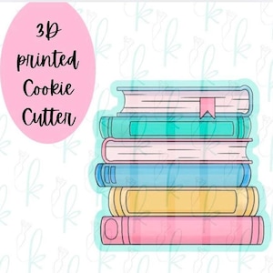 May include: A 3D printed cookie cutter shaped like a stack of five books. The books are coloured in light pink, teal, light blue, yellow, and pink. A pink oval on the left contains the black text 3D printed Cookie Cutter. The design has a light blue outline.