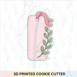 May include: A 3D printed cookie cutter in the shape of a rectangular tag with rounded corners. The design features a small hole at the top with a pink ribbon loop and a green leafy branch extending along the right side of the tag frame.