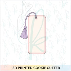 May include: A rectangular cookie cutter with rounded corners, featuring a light pink center and a darker pink border. A lavender-colored string is attached to the top, ending in a matching lavender tassel. The text 3D PRINTED COOKIE CUTTER is visible.