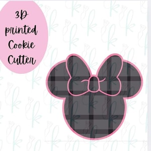 May include: A 3D printed cookie cutter shaped like a mouse head with a large bow. The design features a dark gray and black plaid pattern with a bright pink outline. The text 3D printed Cookie Cutter is displayed in a pink oval on the left side.