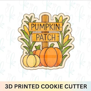 May include: A 3D printed cookie cutter in the shape of a pumpkin patch sign. The sign reads "Pumpkin Patch" and is flanked by corn stalks. Three pumpkins are at the base of the sign, with a light yellow pumpkin on the left and two orange pumpkins on the right.