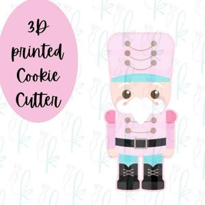 May include: A 3D printed cookie cutter shaped like a nutcracker. The design features a light pink uniform with a black belt, dark boots, and a white beard. To the left, a pink oval contains the black text 3D printed Cookie Cutter.