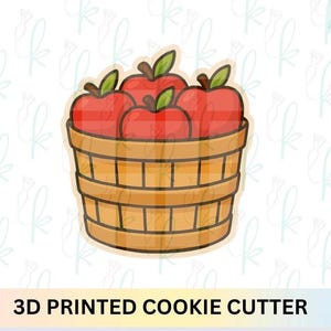 Puede incluir: Un cortador de galletas impreso en 3D que presenta una cesta llena de cuatro manzanas rojas. Las manzanas tienen hojas verdes y tallos marrones. La cesta es de madera marrón claro con listones verticales y horizontales.