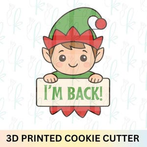 May include: A 3D printed cookie cutter featuring a cartoon elf holding a sign that reads "I'M BACK!". The elf has a green hat with a red pom-pom, a red collar, and a smiling face. The text "3D PRINTED COOKIE CUTTER" is at the bottom.