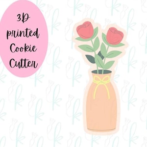 May include: A 3D printed cookie cutter shaped like two pink roses in a peach-colored vase with a yellow ribbon tied around the neck. The text 3D printed Cookie Cutter is displayed in a pink oval on the left side of the image.