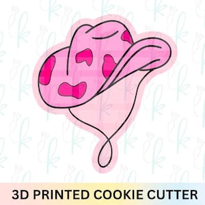 May include: A pink 3D printed cookie cutter in the shape of a cowboy hat with dark pink spots. The hat has a black outline and a curved line extending from the bottom. The text "3D PRINTED COOKIE CUTTER" is at the bottom.